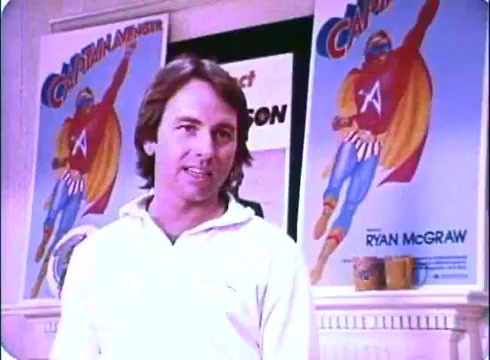 The still from the 'Hero at Large' movie promo features John Ritter in a white shirt, standing in front of two colorful posters of a superhero character named Captain Avenger. The posters prominently display the superhero in a dynamic pose, suggesting action and excitement. The backdrop includes elements indicating a promotional setting, creating a lighthearted tone typical of the film's comedy genre. This scene encapsulates Ritter's charismatic presence as he engages with the viewer.