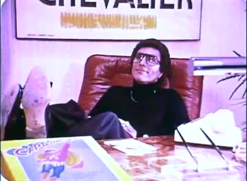 The still from the 'Hero at Large' movie promos features a character reclining in an office setting, leaning back in a chair with his feet propped up on a desk. He is dressed in a black turtleneck and has glasses, conveying a casual yet confident demeanor. The background includes a poster with the word 'CHEVALIER' and various items on the desk, including a comic book titled 'Capitan', hinting at a lighthearted, comedic atmosphere. The overall aesthetic reflects the style of early 1980s cinema.