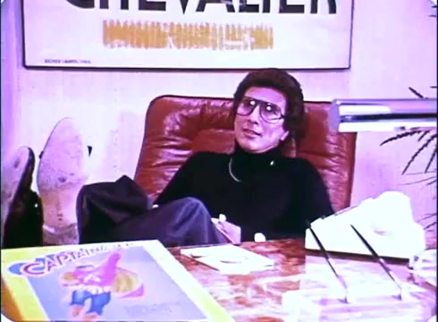 The still from the 'Hero at Large' movie promo features a character seated in an office setting, casually reclining with feet up on a desk. The individual wears glasses and a black outfit, suggesting a laid-back attitude. The desk is cluttered with various items, including a magazine or promotional material featuring a colorful cover. A poster in the background adds some visual interest, hinting at the film's comic or lighthearted nature. The overall vibe reflects the 1980s style of comedy portrayed in the movie.