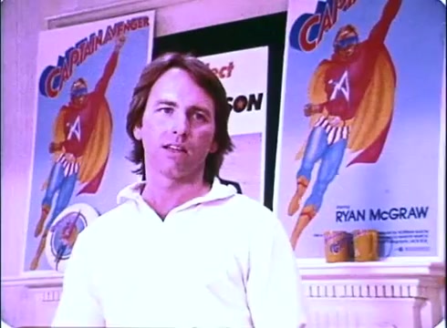 The still features John Ritter in a casual white shirt, standing in front of vibrant promotional posters for the fictional superhero 'Captain Avenger.' The posters depict a heroic character in a colorful costume, reinforcing the comedic superhero theme of the 1980 film 'Hero at Large.' The scene conveys the lighthearted and playful tone of the movie, highlighting Ritter's role in a story about an everyday man becoming a superhero.