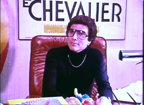 The still from the 'Hero at Large' movie promo features a man with curly hair and large glasses, dressed in a black turtleneck. He is seated at a desk cluttered with colorful toys and papers, exuding a retro vibe typical of the early 1980s. Behind him is a poster that reads 'LE CHEVALIER,' setting the scene in an office environment, likely related to the film's comedic and playful tone. The overall aesthetic reflects the time period and is indicative of the film's lighthearted nature.