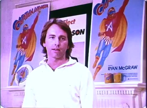 The still features John Ritter in a white sweater, standing in a room decorated with promotional posters for a superhero character, 'Captain Avenger.' The film, 'Hero at Large,' combines comedy with themes of heroism. The bright colors of the posters contrast with the more subdued tones of the setting, creating a playful atmosphere that reflects the film's lighthearted nature. Ritter's expression suggests a blend of humor and sincerity, characteristic of his performance in the movie.
