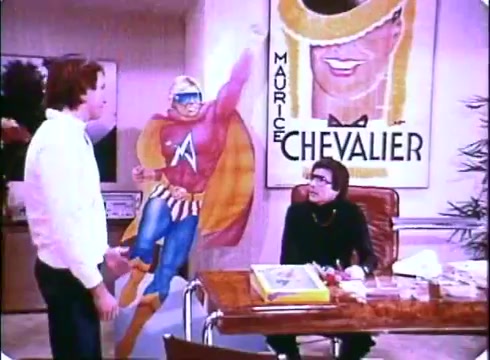 The still from 'Hero at Large' features two characters in an office setting. One character, dressed in a superhero costume, is depicted as a life-sized cutout, while the other character, presumably John Ritter, is engaging in conversation with a seated figure at a desk. The seated figure appears to be dressed in a formal outfit and is looking at the other character. Behind them, a vibrant poster of Maurice Chevalier adds a colorful backdrop to the scene. The overall aesthetic is characteristic of late 1970s to early 1980s cinema, with a focus on humor and whimsy in the context of a comedy film.