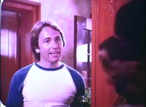 The still from 'Hero at Large' features John Ritter in a casual outfit, wearing a white t-shirt with blue sleeves. He appears to be engaged in a conversation, likely at the doorway of an interior setting. The aesthetic reflects the film's comedic tone, and the color dynamics suggest a warm, inviting atmosphere typical of the late 1970s and early 1980s. This scene promotes the film's light-hearted approach to storytelling.
