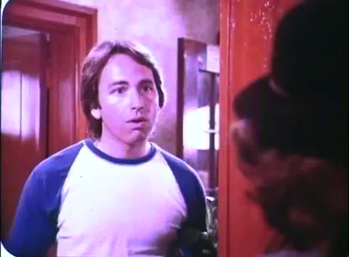 The still from 'Hero at Large' features a young man with shoulder-length brown hair, wearing a blue and white raglan tee, standing in an interior setting. He appears surprised or concerned as he looks towards another character off-camera. The background suggests a domestic scene, with warm lighting that evokes a casual atmosphere. The film's comedic tone is likely reflected in this moment of interaction.