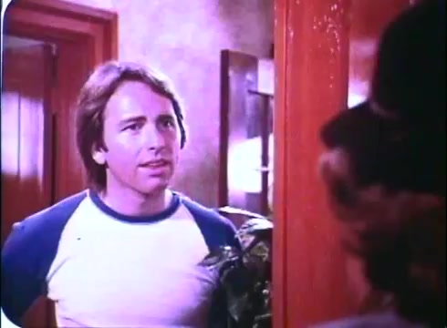 The still from 'Hero at Large' features John Ritter in a casual outfit, specifically a white shirt with blue sleeves. He appears to be engaged in a conversation, likely surprised or intrigued, as he stands in a doorway, framed by wooden panels and light from behind him. The setting suggests an interior scene, possibly at a home, and there's a hint of greenery nearby. The overall tone captures a moment of interaction, characteristic of the film's comedic style.