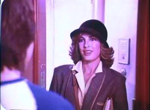 The still from the 'Hero at Large' movie trailer features a woman, likely a character in the film, wearing a stylish hat and a blazer. She appears to be greeting another character, who is mostly out of view. The setting suggests a warm or lighthearted moment, fitting for the comedy genre. The color tone reflects typical film aesthetics of the late 1970s and early 1980s.