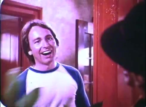 The still from 'Hero at Large' features John Ritter with a bright smile, wearing a casual blue and white shirt. He appears to be in a doorway, engaging with someone outside of the frame. The background suggests a home setting, with warm tones and wooden decor. The overall mood conveys a sense of friendliness and humor, fitting the comedic tone of the film.