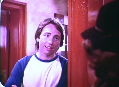 The still features John Ritter as the character in 'Hero at Large,' dressed in a casual white and blue shirt. He appears to be engaged in a conversation, likely at home, with a doorway framing his figure. The setting includes warm, earthy tones, with wooden elements typical of the early 1980s aesthetic. The scene conveys a light-hearted, comedic atmosphere, reflective of the film's themes.