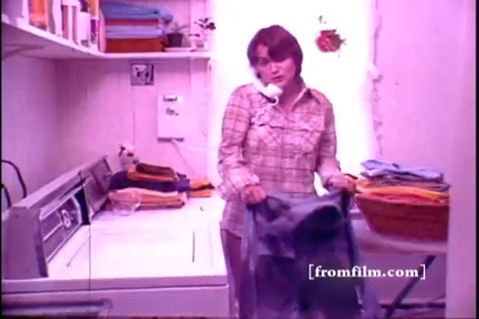 The still features a woman in a laundry room, holding a pair of jeans while talking on the phone. She has shoulder-length brown hair and wears a plaid shirt, conveying a casual, domestic setting. The room is filled with stacks of colorful towels and a washing machine, with a soft pink hue indicating the vintage 16mm print quality. The scene reflects a typical 1970s household chore, likely promoting Lestoil detergent.