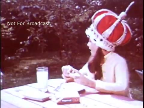 The still features a woman wearing a large, ornate crown, sitting at an outdoor picnic table. She is holding a sandwich in one hand while looking away. A glass of milk sits on the table next to her, along with a box of Imperial Margarine. The background is a greenery, suggesting a casual outdoor setting typical of 1970s advertisements. The overall tone conveys a playful and lighthearted vibe associated with the brand.