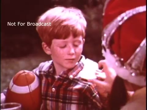 The still features a young boy with reddish hair, wearing a plaid shirt, sitting outdoors. He looks pleasantly surprised as he interacts with a person wearing a festive costume, likely a holiday character. A foam football is in his lap, and there is a drink beside him, suggesting a casual, cheerful gathering, typical of a children's commercial from the 1970s promoting Imperial Margarine.