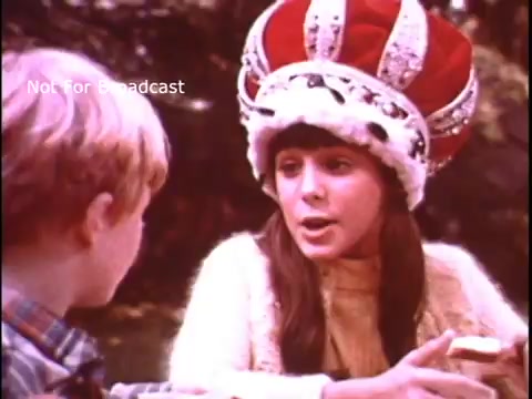 In the 16mm still from the 1970s Imperial Margarine commercial, a young girl wearing a large, ornate crown speaks to a boy. She appears to be sharing a message about the product, with a joyful expression, while holding a piece of bread with margarine on it. The scene is set in a natural outdoor environment, reflecting a playful and whimsical atmosphere typical of that era's advertisements.