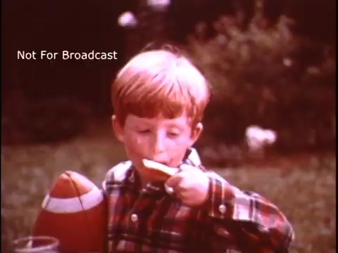 The still features a young boy with red hair, wearing a plaid shirt, seated outdoors. He holds a slice of bread spread with margarine and is likely enjoying a snack. A toy football is tucked under his arm, and the background is blurred with greenery, hinting at a sunny day. The scene conveys a sense of nostalgia and simplicity, typical of 1970s advertisements. The text 'Not For Broadcast' is superimposed.
