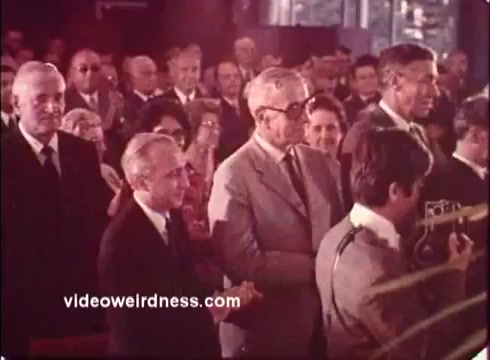 The still from the 'Blue Bonnet Margarine Commercial - 1970s' features a group of people gathered in what appears to be an indoor event or gathering. The atmosphere is formal, with attendees dressed in suits. Some individuals are applauding, while others are engaged in conversation or posing for a camera. The color palette reflects the typical hues of the era, adding a nostalgic feel. The image captures a moment that suggests celebration or recognition, possibly related to the product being promoted.   For more content, follow on Twitter @FromFilm or like and subscribe on YouTube.