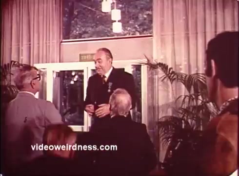 This still from a 1970s Blue Bonnet Margarine commercial depicts a scene in a brightly lit indoor setting, likely a gathering or event. The image shows several individuals, including a prominently dressed man at the center, engaging in conversation. The background features large windows with a view of greenery outside and decorative hanging lights. The atmosphere appears cordial and lively, characteristic of promotional marketing from that era.   For more interesting content, follow @FromFilm on Twitter or subscribe on YouTube.