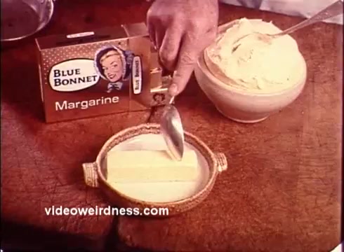 The still from the 1970s Blue Bonnet Margarine commercial features a hand holding a spoon, poised to scoop margarine from a dish. Next to it is a colorful box of Blue Bonnet Margarine, depicting a woman on the packaging. In the foreground, a butter dish holds a block of margarine, while a bowl filled with a creamy mixture sits nearby. The scene reflects a vintage kitchen setting, typical of 1970s advertisements that emphasize the freshness and versatility of the product.
