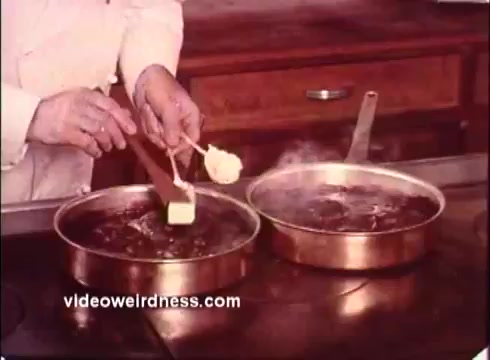 The still from the 'Blue Bonnet Margarine Commercial - 1970s' features a hand in a white chef's coat adding a pat of margarine to two frying pans on a stovetop. One pan is bubbling, indicating heat, while the other one is also being prepared. This imagery highlights the cooking process and showcases Blue Bonnet Margarine as a key ingredient in meal preparation during that era. The warm color tones typical of 16mm film add a nostalgic feel to the scene.