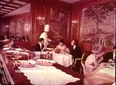 The still from the 1970s Blue Bonnet Margarine commercial showcases a formal dining setting, likely in a restaurant. It features elegantly dressed patrons at tables with meals displayed, while a waiter in a chef's hat serves food. The wooden paneling and decor exude a classic charm, reflecting the visual style of that era. The overall vibe suggests an emphasis on fine dining and quality, in line with the promotional message of the margarine product.