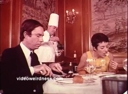 The still from the 'Blue Bonnet Margarine Commercial - 1970s' features a formal dining setting where a man, dressed in a suit, is engaged in his meal, slicing food on his plate. At the same table, a woman in a bright yellow dress is enjoying her meal as well. In the background, a chef in a white coat and tall hat is seen pouring something from a jug, possibly adding an element of culinary flair to the scene. The decor reflects the classic style of the era, with elegant furnishings and a warm ambiance, characteristic of promotional materials from that time.