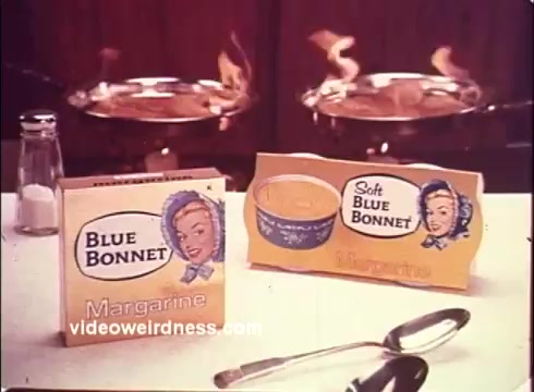 The still from the 1970s 'Blue Bonnet Margarine' commercial features product placement of Blue Bonnet Margarine prominently displayed on a kitchen countertop. The packaging includes a box of margarine and a container labeled 'Soft Blue Bonnet.' The background suggests a cooking environment, possibly with pots or pans that may be in use, indicated by the dynamic visual of flames. The overall aesthetic captures the vintage design and marketing style of the era, aimed at showcasing the appeal of the margarine product.