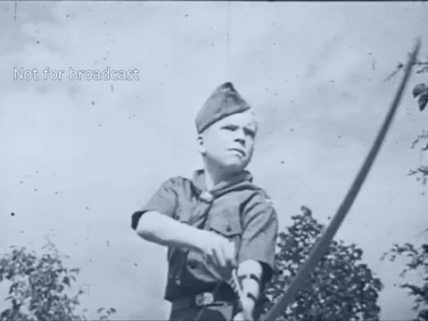 The still features a young boy, dressed in a Boy Scout uniform with a hat, holding a bow with an arrow drawn back, ready to shoot. The background shows trees and a clear sky, suggesting an outdoor scouting activity. The image captures a moment of focus and determination, typical of a public service announcement promoting scouting skills in the 1960s. A watermark indicates that it's not meant for broadcast.