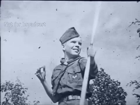 The still features a young boy in a Boy Scout uniform, smiling and holding a stick, possibly representing a scouting tool or flag. The background includes trees, suggesting an outdoor setting typical of scouting activities. The image conveys a sense of adventure and camaraderie associated with scouting life in the 1960s.