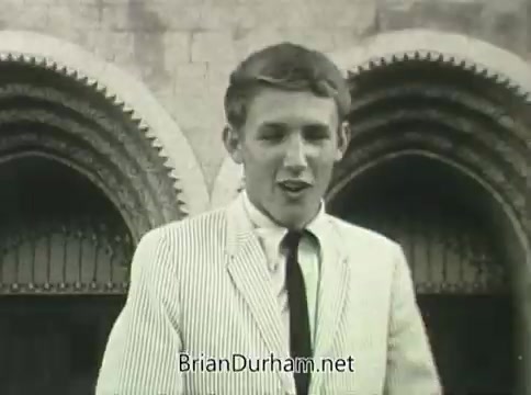 The still from the 1968 Ford 'Miss Teenage America' PSA depicts a young man in a striped suit and tie, standing in front of an architectural structure with arches. His expression suggests enthusiasm, fitting the upbeat theme of the PSA, which likely promotes self-confidence among teenagers. The image shows signs of vinegar syndrome, resulting in visible warping and degradation, reflecting its age and condition.