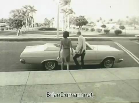 The still from the 'Ford Miss Teenage America' PSA (1968) features a young couple standing next to a classic convertible car. The scene is set in a sunny, suburban environment, with palm trees visible in the background. The film appears warped due to vinegar syndrome, affecting its clarity and visual quality. The couple's attire reflects the fashion of the era, contributing to the nostalgic aesthetic of the PSA.