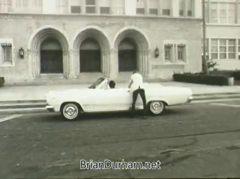 The still from the 1968 Ford 'Miss Teenage America' PSA depicts a white convertible parked on a street in front of a building with architectural details. A figure stands by the car, possibly interacting with someone inside. The image is affected by vinegar syndrome, resulting in visual distortion and warping. The overall aesthetic reflects the style of the late 1960s, highlighting themes of youth and Americana.