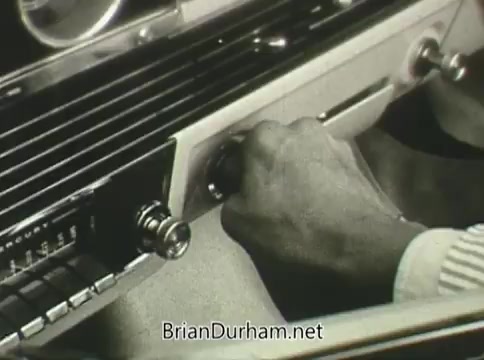 The still from the 1968 Ford 'Miss Teenage America' PSA shows a close-up of a hand adjusting the radio in a classic car. The image exhibits signs of vinegar syndrome, indicating deterioration, characterized by warping and discoloration. The focus is on the vintage car interior, emphasizing the era’s design and technology.