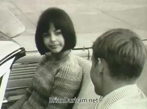 The still from the 1968 PSA 'Miss Teenage America' showcases a young girl with a bob haircut in a car, engaging with a young man. The image conveys a sense of youth and interaction typical of the era, but the quality is affected by vinegar syndrome, giving it a distorted, aged appearance. The scene captures the cultural context of the time, focusing on teenage life and automotive interests.