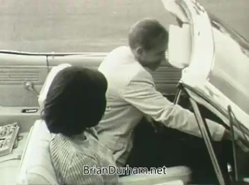 The still from the Ford 'Miss Teenage America' PSA from 1968 features a young woman and a man seated in a convertible car. They appear engaged in conversation, with the man reaching towards the dashboard. The image may showcase a mid-20th century style, reflective of the era's automotive designs. Notably, the film exhibits signs of vinegar syndrome, resulting in a distorted and warped appearance.