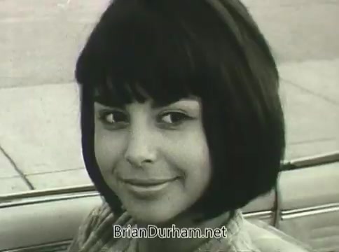 The still from the 1968 Ford 'Miss Teenage America' PSA features a young girl with a bob haircut, smiling warmly at the camera. The image has a vintage aesthetic, characteristic of the era, and shows signs of deterioration due to vinegar syndrome, giving it a warped and aged appearance. The background appears to be a street scene, but details are muted due to the film's condition.