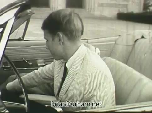 The still from the 1968 PSA 'Ford Miss Teenage America' features a man in a light-colored striped suit, seated in the driver’s seat of a car. The background suggests a classic or upscale setting with visible architecture. The image shows signs of deterioration consistent with vinegar syndrome, such as warping and color fading, which gives the still a nostalgic and vintage feel. The overall tone reflects the era’s style and societal context surrounding teenage culture and automotive branding.