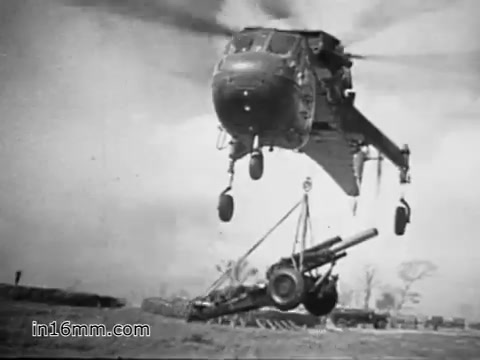 The still from 'Your Man in Vietnam' (1968) features a CH-54 helicopter lifting artillery while flying above a field. The scene highlights the helicopter's utility in transporting heavy equipment during the Vietnam War, showcasing its role and the efforts of the U.S. Army personnel, specifically Specialist 6 James Daly, who operates and maintains the aircraft. The image captures the ruggedness of military aviation in conflict zones.