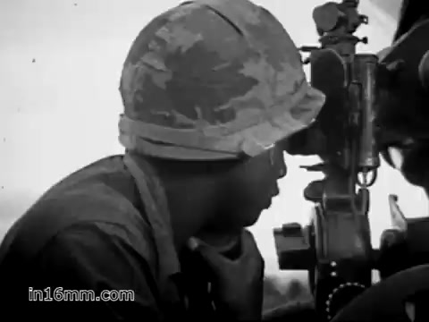 The still depicts a soldier, likely of African descent, wearing a camouflage helmet and focused intently on a weapon, possibly a machine gun. His expression conveys concentration and determination. The background suggests a war setting, emphasizing the intensity of military life during the Vietnam War era. The image reflects the challenges faced by soldiers, highlighting their roles and responsibilities in combat situations.