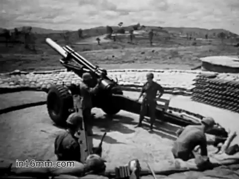 The 16mm still from 'Your Man in Vietnam' features a group of soldiers operating a large artillery piece, likely illustrating the logistical aspects of military operations during the Vietnam War. The composition shows the soldiers in a sandbagged position, highlighting the rugged terrain typical of the Vietnam landscape. The scene emphasizes the heavy machinery used in warfare, reflecting the message of the film about the various roles soldiers had to take on, including operating and maintaining such equipment.