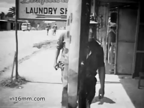The still from 'Your Man in Vietnam' (1968) depicts a soldier, presumably Specialist 6 James Daly, walking past a laundry shop in a rural Vietnamese setting. He appears focused, with military attire suggesting his role in the U.S. Army during the Vietnam War. The background shows a dusty street, typical of the era, with civilians and other soldiers in the distance, reflecting the daily life and atmosphere of that time.