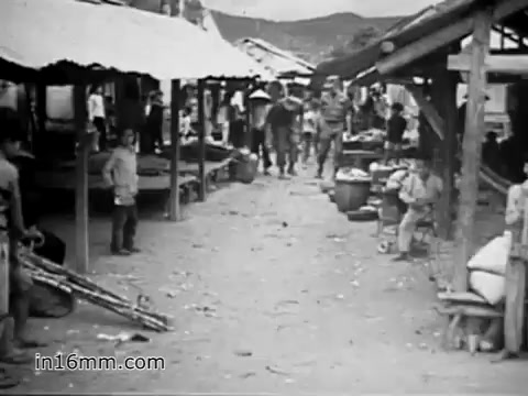 The still from 'Your Man in Vietnam' captures a bustling marketplace scene, likely in a rural area of Vietnam during the late 1960s. The setting features simple structures with thatched roofs, with people moving about—some interacting, others engaged in trade. Children are visible, adding a sense of everyday life amidst the backdrop of military presence hinted at in the voiceover. This environment reflects the contrast between the civilian world and military operations described by James Daly, a flight engineer managing the CH-54 helicopter.