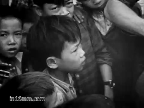 The still captures a moment from the 1968 U.S. Army recruiting film 'Your Man in Vietnam.' It features a group of Vietnamese children, with a focus on a boy in the foreground. He looks contemplative, surrounded by peers who gaze intently at something off-camera. The image reflects the interplay between the U.S. military presence and the local population during the Vietnam War, emphasizing the poignant and complex interactions that defined that era.