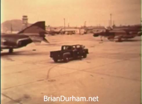 The 16mm still from the 1968 USAF Aerospace ROTC PSA depicts a bustling airfield scene. In the foreground, a vintage pickup truck is parked on the tarmac, while in the background, several military aircraft, likely fighter jets, are lined up. The overall atmosphere reflects a sense of teamwork and dedication to service, embodying the spirit of the Air Force as a path for further education and career development for young men.
