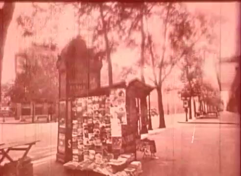The still from 'Erik Satie and the King of the Beans - Part I' features a vintage scene depicting a newsstand adorned with various posters and announcements. The surrounding area is tree-lined, suggesting an urban setting, likely in Paris, reflecting the early 20th-century aesthetic. The image is presented in a sepia tone, adding to the nostalgic and historical context of the film, which explores the life and influence of composer Erik Satie. The overall composition conveys a sense of stillness and reflection, typical of educational films focused on cultural figures.