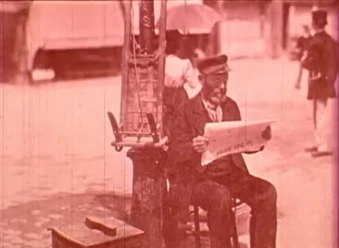 The still from 'Erik Satie and the King of the Beans - Part I' features a man sitting on a wooden chair, intently reading a newspaper. He is dressed in period attire, wearing a cap. In the background, there are hints of a bustling street scene, with blurred figures and an umbrella, suggesting a lively, outdoor setting. The vintage aesthetic is marked by a warm, sepia tone, characteristic of 16mm film, adding a nostalgic quality to the scene.