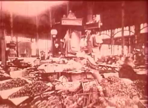 The still from 'Erik Satie and the King of the Beans - Part I' depicts a bustling market scene. Overflowing tables are laden with an array of beans and produce, while vendors and shoppers navigate the lively space. The atmosphere is vibrant and dynamic, highlighted by the architectural details of the market structure, with large windows allowing natural light to fill the area. The overall tone is warm and nostalgic, reflecting the essence of a community gathering place.
