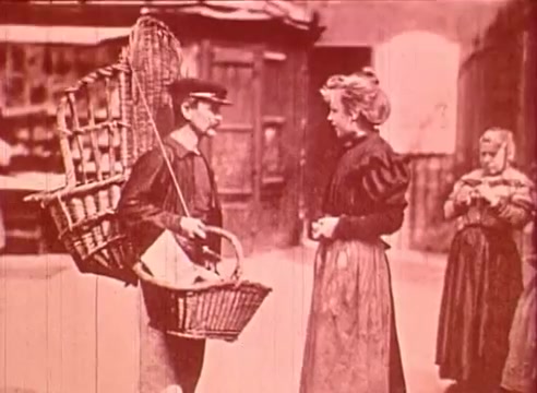 The still from 'Erik Satie and the King of the Beans - Part I' features a scene set in a vintage urban environment. A young man, dressed in period attire and carrying a large basket of goods, engages in conversation with a woman in a long skirt and blouse. She appears to be interested or intrigued by the items he is selling. A young girl stands to the side, observing the interaction. The image conveys a nostalgic atmosphere, reflecting the everyday life and commerce of the time.