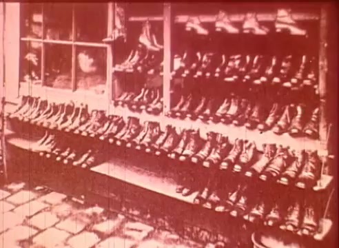 The still from 'Erik Satie and the King of the Beans - Part I' features a display of numerous boots lined up on shelves. The image has a vintage appearance, likely tinted in sepia tones, reflecting the early film aesthetics. The setting suggests a shop or showroom, emphasizing the organized arrangement of the footwear. This scene may serve an educational purpose, potentially illustrating themes related to commerce, fashion, or Satie's cultural context.