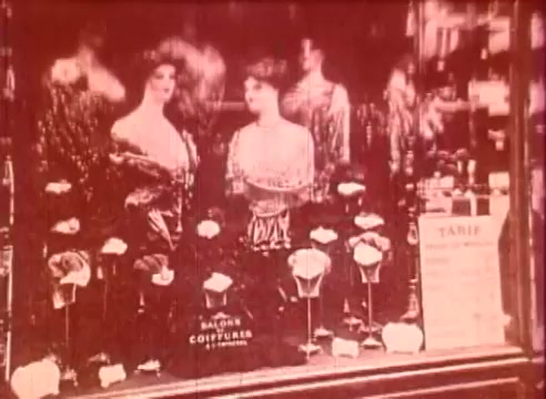 The still from 'Erik Satie and the King of the Beans - Part I' features two elegantly dressed women displayed in a shop window. They are surrounded by roses and other decorative elements, suggesting a vintage fashion salon. The warm tones and soft focus evoke a nostalgic atmosphere, typical of early 20th-century aesthetics, aligning with the film's educational themes related to art and culture.