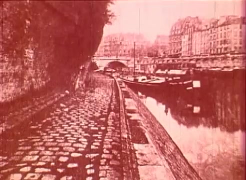 The still from 'Erik Satie and the King of the Beans - Part I' features a scenic view along a cobblestone pathway beside a river. On one side, there is a stone wall, while the opposite side shows moored boats reflecting in the water. Historic buildings line the background, contributing to a nostalgic ambiance, all presented in a warm, reddish tone. The frame encapsulates an educational perspective on the artistic environment surrounding Erik Satie.
