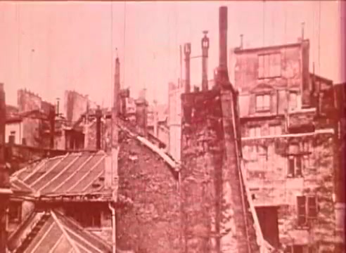 The still from 'Erik Satie and the King of the Beans - Part I' features a sepia-toned cityscape, showcasing old buildings and rooftops in a somewhat dilapidated state. The architecture suggests an early 20th-century urban environment, with chimney stacks and varied rooflines. This visual context relates to the educational aspect of the film, likely aiming to evoke the historical backdrop of Erik Satie's era.